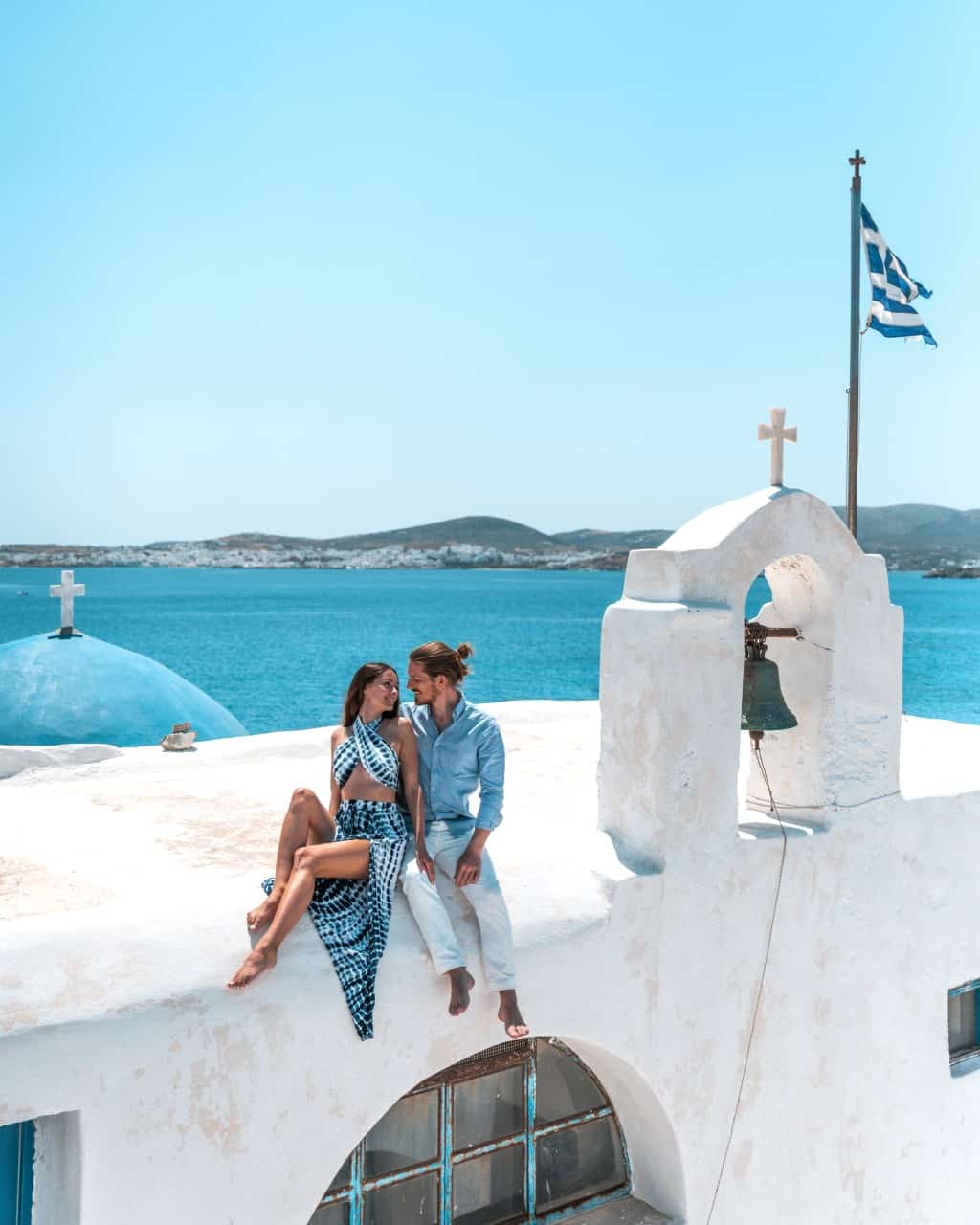 A photo of us sitting at the white-washed Monastery of St. John Deti in Paros, with its vibrant blue dome, a waving Greek flag, and the turqouise sea in the background.