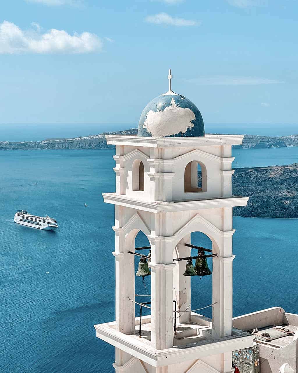 A white bell tower with a blue dome overlooking the Aegean Sea, with a boat and a few islets in the background.