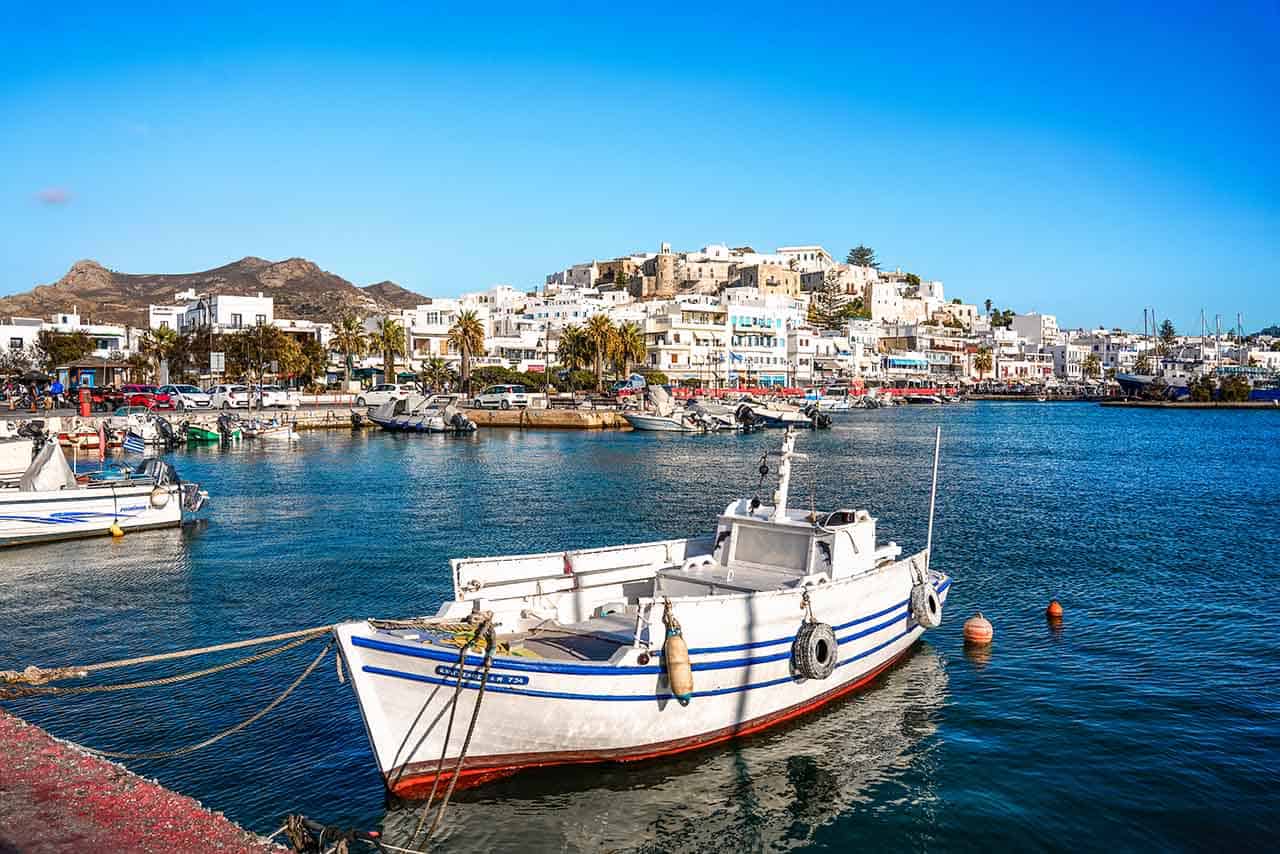 A scenic view of Naxos Town and the harbor, featuring fishing boats anchoring in the water on a sunny day
