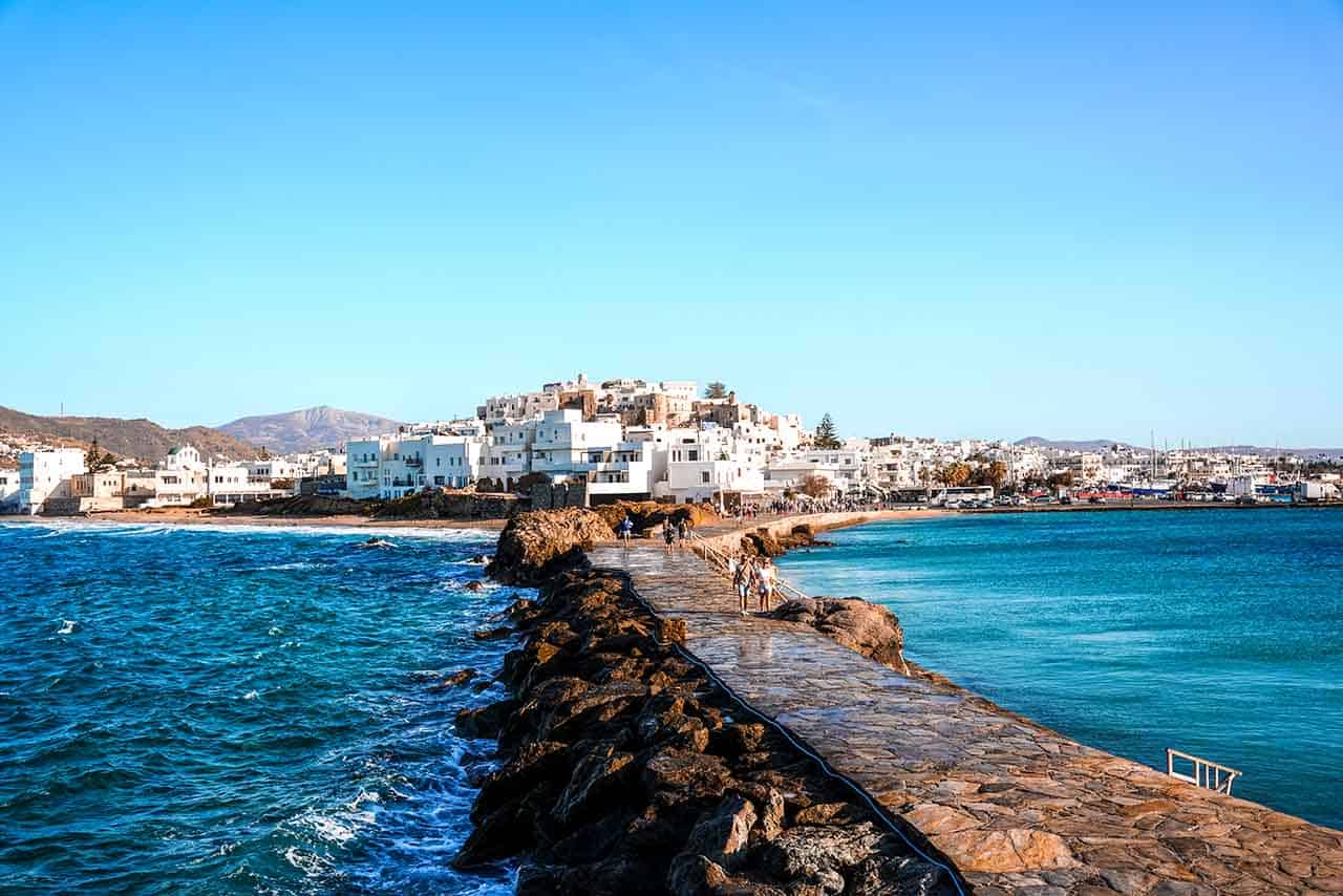 A scenic view of Naxos Town, featuring a stone walkway leading to the heart of the town, with blue, wavy waters flanking both sides.