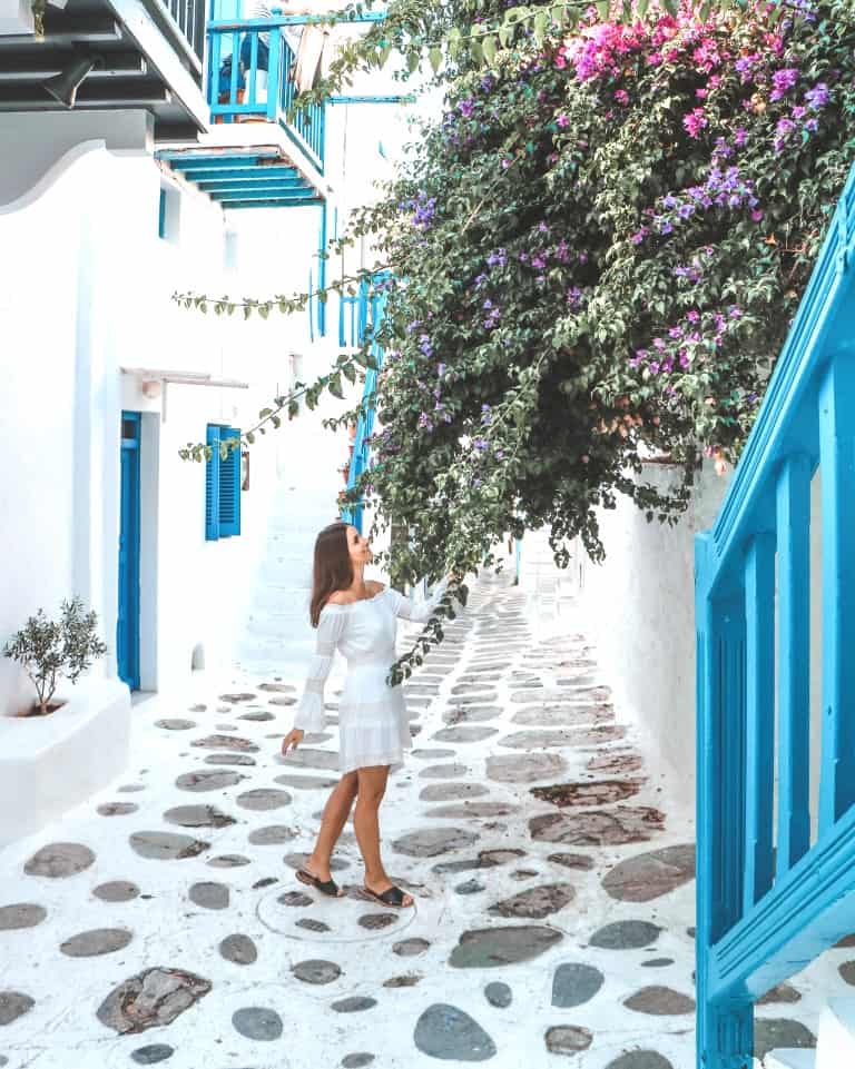 A photo of me standing beside a vibrant bougainvillea tree, nestled in a picturesque cobblestoned alley adorned with white houses and accented by blue doors, windows, and stairs.