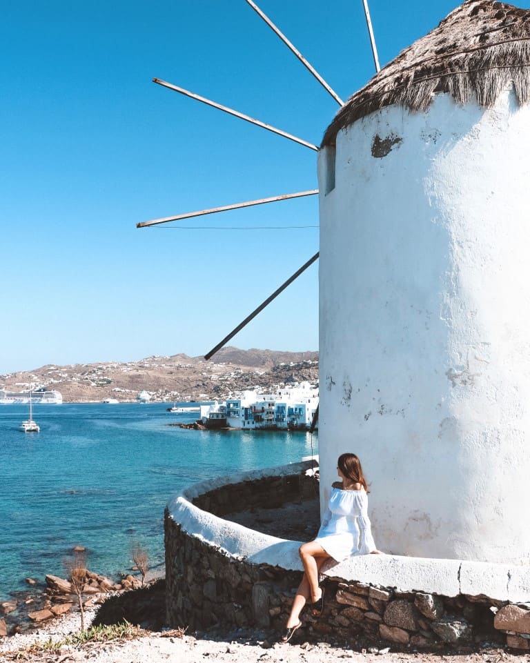 A photo of me seated in front of a windmill in Mykonos, with the deep blue sea and the picturesque Mykonos Town as a scenic backdrop.