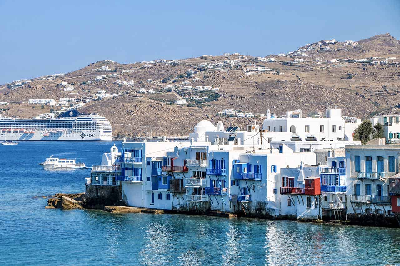 A photo of Little Venice in Mykonos, where charming white-washed houses line the seafront, with a big cruise ship and scenic hills in the background.