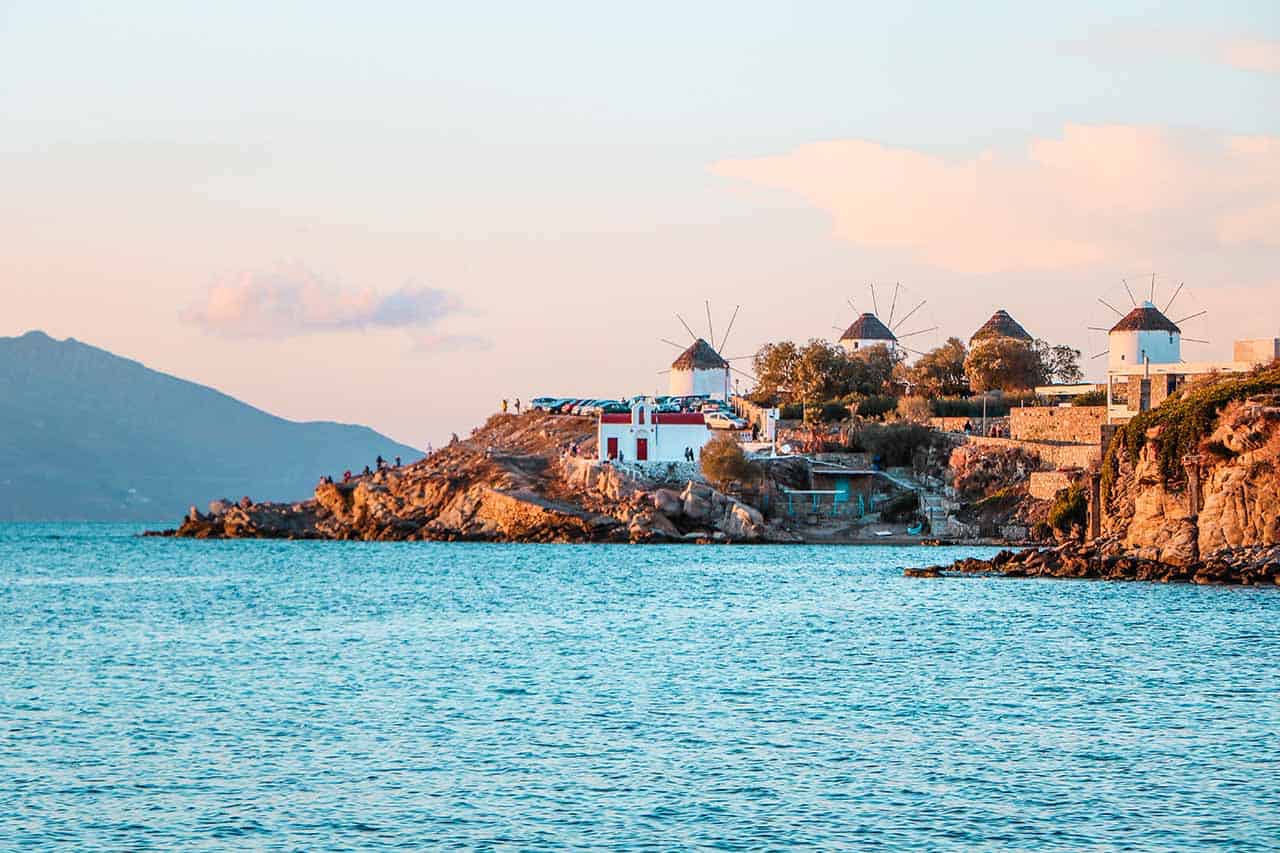 A dreamy image featuring four traditional Cycladic windmills and a small chapel perched along the coastline, against a backdrop of a pastel-colored sky.