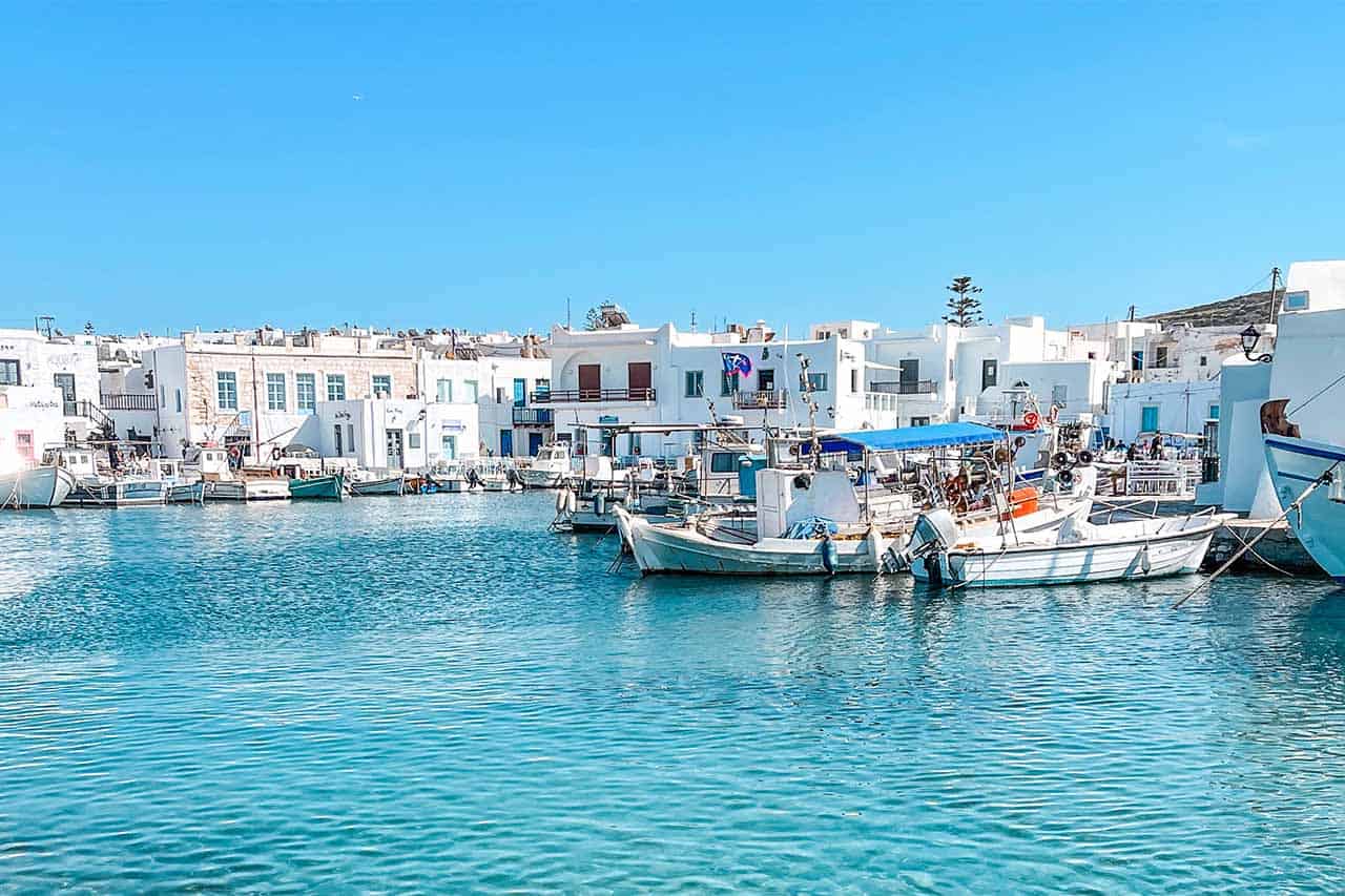 A photo of the charming old harbor in Paros, where small fishing boats dot the turquoise waters, framed by white-washed houses under a clear blue sky.